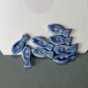 May include: A collection of eight ceramic fish-shaped buttons. The buttons are primarily blue with white accents, featuring two holes for sewing. These decorative buttons are ideal for sewing and craft projects.