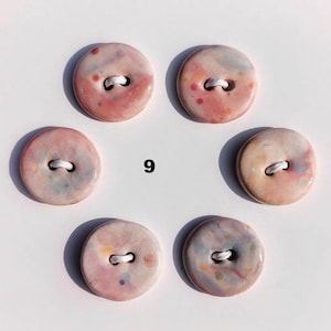 May include: Six round ceramic buttons with a speckled pink, peach, and blue design. Each button has a white thread for sewing. The number "9" is printed in black between two buttons. These buttons are ideal for sewing and craft projects.