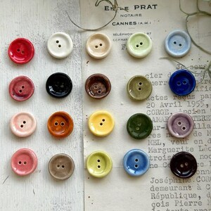 Könnte beinhalten: Eine Sammlung von zwanzig runden Keramikknöpfen, die in einem Raster angeordnet sind und eine Vielzahl glänzender Farben aufweisen, darunter Rot, Weiß, Beige, Hellgrün, Blau, Schwarz, Braun, Rosa, Orange, Gelb, Dunkelgrün, Lila und Dunkelbraun, jeweils mit zwei Löchern.