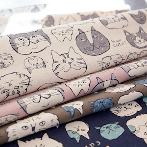 May include: Several fabric rolls with cat-themed patterns. The designs feature various cat faces in black and gray on a light beige background. Other fabrics have pink, brown, and navy blue backgrounds with similar cat illustrations. Some fabrics have text that says "How cute!" and "lovely cat".