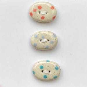 Peut inclure: Trois boutons en céramique avec un glaçage blanc cassé moucheté. Chaque bouton a des pois de couleurs différentes : rouge, bleu clair et sarcelle. Les boutons ont deux trous pour la couture.