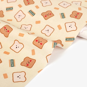 May include: A cream-colored fabric featuring a repeating pattern of cartoon toast slices with simple facial expressions, some with pink cheeks, interspersed with small rectangular butter sticks labeled with the word BUTTER in green text.