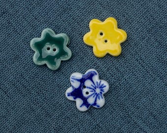 May include: Three ceramic flower-shaped buttons. One is green, one is yellow, and one is white with blue and yellow floral designs.