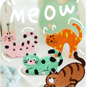 Könnte beinhalten: Vier gestickte Bügelbilder mit niedlichen Comic-Katzen in verschiedenen Posen. Die Katzen sind rosa mit schwarzen Flecken, orange mit gelben Flecken, grün mit schwarzen Flecken und braun mit orangefarbenen Streifen. Das Wort "Miau" ist in Weiß über den Patches geschrieben.