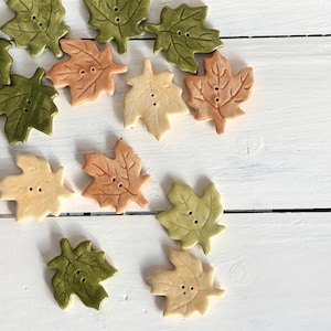 May include: A collection of ceramic buttons shaped like maple leaves in shades of olive green, sage green, and light tan. Each button features a detailed leaf vein texture and two central holes for sewing, arranged in a scattered pattern.