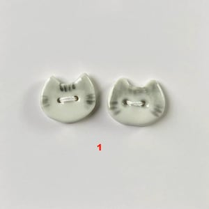 Può includere: Due bottoni in ceramica a forma di gatto. I bottoni sono bianchi con strisce grigie e dettagli del viso. Ogni bottone ha due fori per cucire. Il numero "1" è stampato in rosso sotto i bottoni.