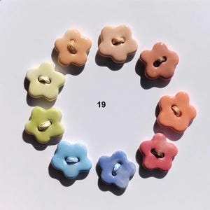 May include: A collection of nine flower-shaped buttons arranged in a circle, each featuring a glossy finish and a central hole with thread. The colors include shades of peach, pink, yellow, green, and blue. The number 19 is printed in the center.