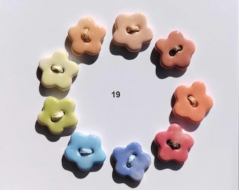 Könnte beinhalten: Eine Sammlung von neun blumenförmigen Knöpfen, die in einem Kreis angeordnet sind. Jeder Knopf hat eine glänzende Oberfläche und ein zentrales Loch mit Faden. Die Farben umfassen Pfirsich, Rosa, Gelb, Grün und Blau. Die Zahl 19 steht in der Mitte.