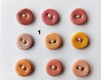 May include: A grid of nine round, smooth buttons arranged in three rows. The buttons feature various shades of pink, orange, and yellow, each with a central hole containing a light-colored thread. The number 1 is printed in black near the center.