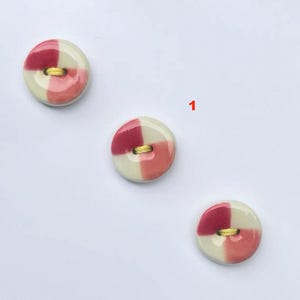 以下が含まれることがあります： クリーム、ピンク、赤の市松模様の丸いボタンが3つ。各ボタンには縫製用の黄色の糸が付いています。数字の「1」が右上隅にあります。