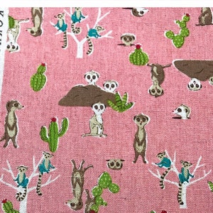 May include: Pink fabric with a repeating pattern of meerkats, cacti, and trees. The meerkats are in various poses, including standing, sitting, and looking up. The cacti are green and have spines. The trees are white with brown branches. The fabric has a textured surface.