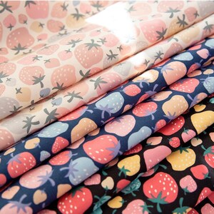 May include: Close-up of several fabric patterns featuring strawberry designs. The fabrics display strawberries in various colors, including pink, red, yellow, and blue, against backgrounds of white, navy, and black. The strawberries have green stems.