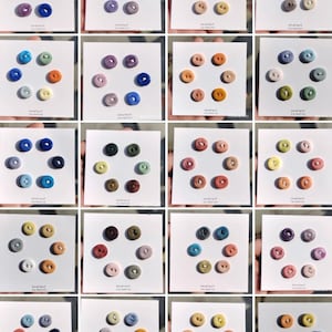Puede incluir: Una colección de pequeños botones de cerámica coloridos dispuestos en patrones circulares sobre tarjetas blancas. Los botones vienen en varios tonos, incluyendo azul, verde, amarillo, naranja y rosa. El texto "smallest ceramist" es visible en cada tarjeta.
