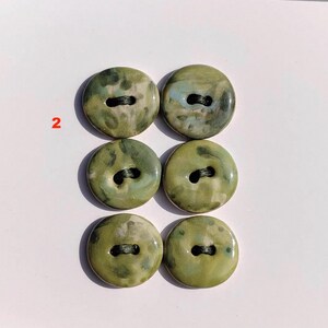 May include: Six round, olive green buttons with black thread. The buttons have a marbled appearance with hints of blue and white. The number "2" is visible in the upper left corner. These buttons are suitable for sewing projects.
