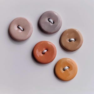 Op de afbeelding: Vijf ronde, glanzende knopen in tinten roze, mauve, oranje en bruin. Elke knoop heeft een witte draad die door twee gaten is genaaid. De knopen zijn gerangschikt op een wit oppervlak.
