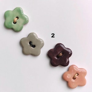 May include: Four ceramic flower-shaped buttons in shades of green, grey, burgundy, and peach. Each button has a glossy finish and a small hole for threading. The number "2" is visible in the background.