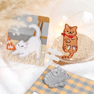 May include: Three embroidered iron-on patches featuring cute cats in different poses. The patches are white, orange, and gray. The white cat is lying down, the orange cat is sitting up with its paws crossed, and the gray cat is curled up.