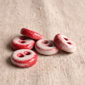 May include: A collection of five round, two-hole buttons featuring a glossy finish with a mottled red and white color pattern. Each button has a smooth, slightly domed surface and a distinct, irregular distribution of red pigment over a white base.