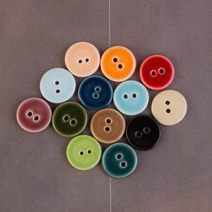Pode incluir: Uma coleção de botões de cerâmica coloridos em várias tonalidades, incluindo pêssego, laranja, vermelho, azul, verde e castanho. Cada botão é redondo com dois furos para costurar. Os botões estão dispostos numa superfície cinzenta.