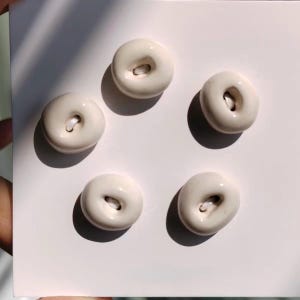 Puede incluir: Cinco botones circulares de color blanco roto, lisos y con acabado brillante, dispuestos en forma de pentágono sobre una tarjeta cuadrada blanca. Cada botón presenta un orificio central con un lazo de hilo blanco visible.