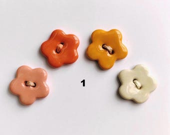 May include: Four flower-shaped buttons in shades of orange, yellow, pink, and cream. Each button has a small hole for sewing. The number "1" is printed in black next to the buttons.