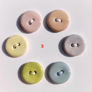 May include: A collection of six ceramic buttons in various pastel colors: pink, peach, yellow, lavender, green, and blue. Each button has two holes and a white thread. The number "3" is printed in red.