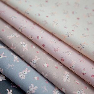May include: Fabric in various pastel shades, including blue, pink, and beige, featuring a repeating pattern of bunnies, cupcakes, and bows. The fabric is folded, showcasing the different colors and patterns.