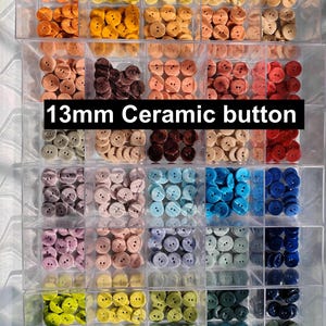 Op de afbeelding: Een verscheidenheid aan 13 mm keramische knopen in een doorzichtige, verdeelde container. De knopen zijn gerangschikt op kleur, waaronder tinten oranje, geel, bruin, roze, blauw en groen. De tekst "13mm Ceramic button" wordt weergegeven.