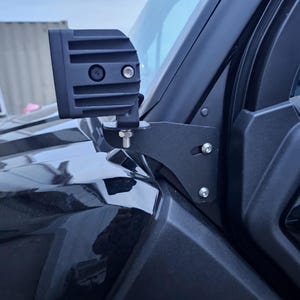 May include: Black LED light bar mounted on a black vehicle. The light bar is attached to the vehicle's frame with a black metal bracket and bolts. The vehicle's windshield and door are visible in the background.