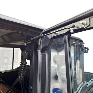 Op de afbeelding: Zwart metalen voertuigdak en deurframe met heldere glazen ramen. Het frame heeft een reeks bouten en scharnieren. Het interieur van het voertuig is zichtbaar, inclusief een stoel en een tas.