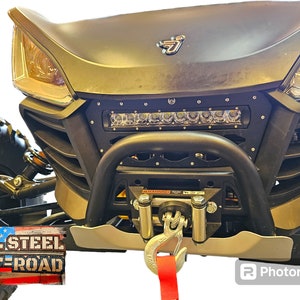 May include: A black and silver off-road vehicle with a black steel bumper and a light bar. The vehicle has a tow hook and a red strap attached to it. The text "ALL STEEL OFF-ROAD" is visible on the lower left side of the image.