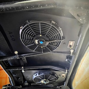 Puede incluir: Dos ventiladores eléctricos negros montados en un marco de metal negro. Los ventiladores son circulares con aspas negras y un centro azul. El marco tiene un interruptor de metal negro en el lado derecho. El texto "ASO" es visible en el lado izquierdo del marco.