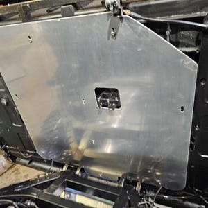 May include: A large, silver metal plate with a rectangular cutout and several screw holes. The plate is mounted on a black surface, possibly a vehicle component. The image is taken from an upward angle.