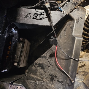 May include: Close-up of a black metal battery box with a red and black wire attached. The box is labeled "ASO" and is mounted to a black metal frame.
