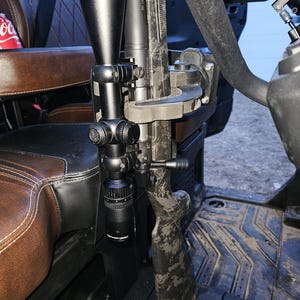 May include: A black rifle with a camouflage pattern is mounted inside a vehicle. A black scope is attached to the rifle. The interior of the vehicle has brown and black leather seats.