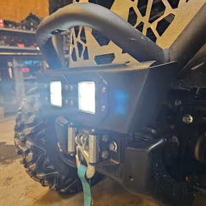 May include: A close-up of a black metal bumper with a set of four bright white LED lights mounted on the front. The bumper is attached to a vehicle with a black tow strap hanging from the bottom.