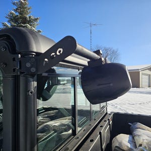 May include: Black speaker system mounted on a black vehicle frame. The speaker is oval-shaped and angled outward. The mounting bracket has cut-out designs. The background shows a snowy landscape and a clear blue sky.