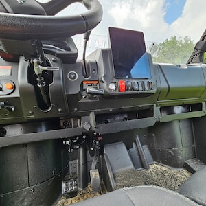 May include: The interior of a black utility vehicle with a black steering wheel, a black dashboard, and a black seat. The dashboard has a touchscreen display, a row of buttons, and a black storage compartment.
