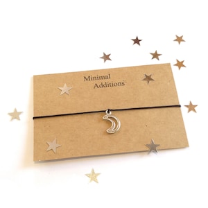 May include: A minimalist bracelet with a silver crescent moon charm on a black cord. The bracelet is presented on a brown card with the text "Minimal Additions". Silver star-shaped confetti is scattered around on a white background.