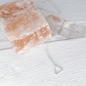 Puede incluir: Un collar de plata con un colgante de tri&aacute;ngulo sobre un fondo blanco. El collar est&aacute; sobre una roca de sal rosa del Himalaya y un cristal de cuarzo transparente.