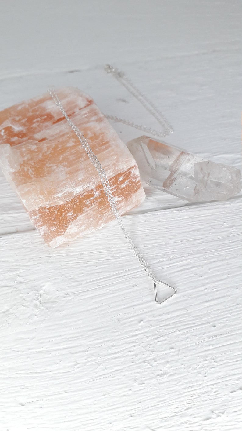 Puede incluir: Un collar de plata con un colgante de tri&aacute;ngulo sobre un fondo blanco. El collar est&aacute; sobre una roca de sal rosa del Himalaya y un cristal de cuarzo transparente.