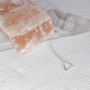 Puede incluir: Un collar de cadena de plata con un peque&ntilde;o colgante triangular. El collar est&aacute; sobre una superficie blanca con una roca de sal rosa y un cristal transparente.