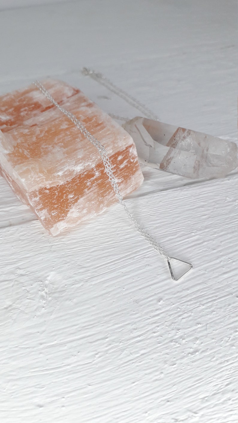 Puede incluir: Un collar de cadena de plata con un peque&ntilde;o colgante triangular. El collar est&aacute; sobre una superficie blanca con una roca de sal rosa y un cristal transparente.