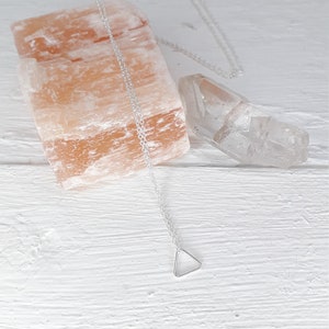 Puede incluir: Un collar de plata con un colgante triangular en una cadena delicada, presentado sobre una superficie de madera blanca con una roca de sal rosa y un cristal de cuarzo transparente.