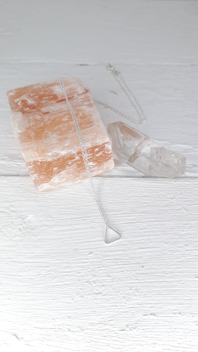 Puede incluir: Un collar de plata con un colgante triangular en una cadena delicada, presentado sobre una superficie de madera blanca con una roca de sal rosa y un cristal de cuarzo transparente.