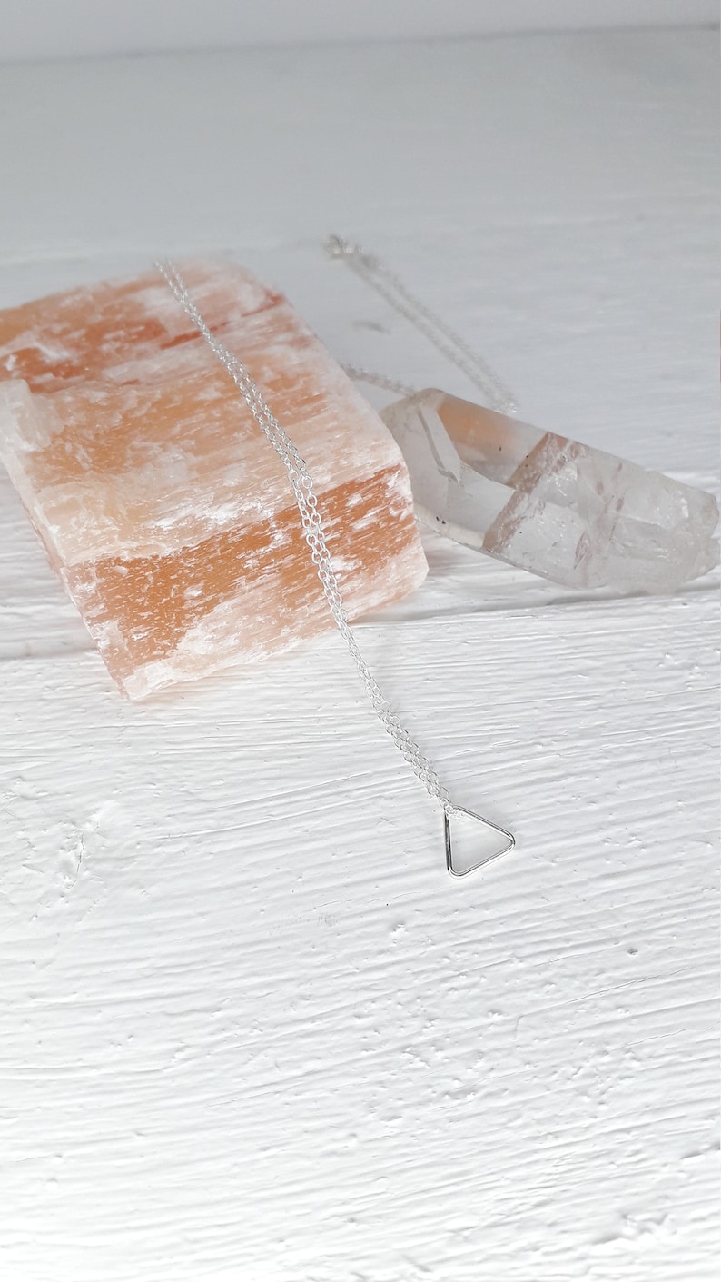 Puede incluir: Un collar de plata con un colgante de tri&aacute;ngulo sobre un fondo blanco. El collar est&aacute; sobre un trozo de sal rosa del Himalaya y un cristal de cuarzo transparente.