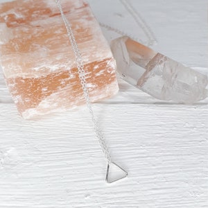 Puede incluir: Un collar de plata con un colgante de tri&aacute;ngulo sobre un fondo blanco. El collar est&aacute; sobre una roca de sal rosa del Himalaya y un cristal de cuarzo transparente.