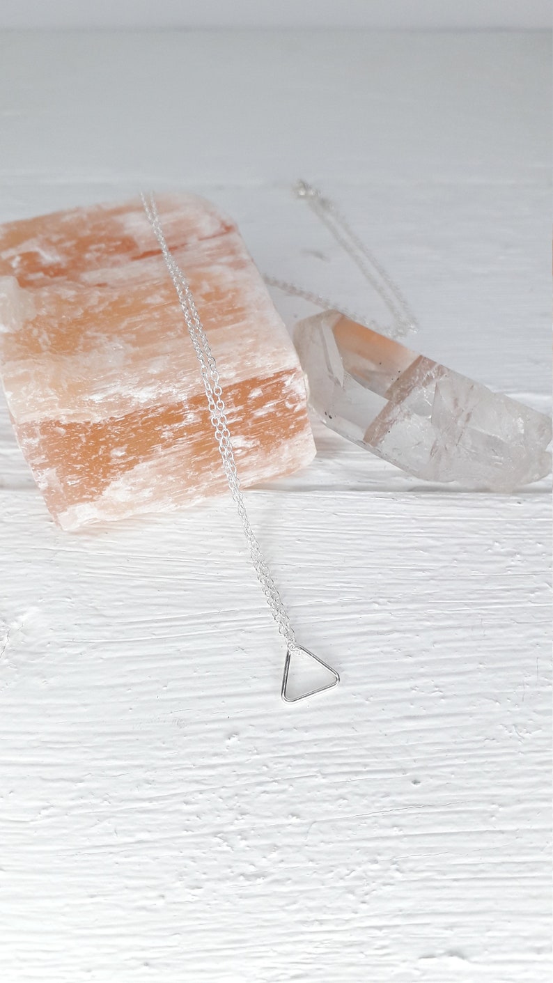 Puede incluir: Un collar de plata con un colgante de tri&aacute;ngulo sobre un fondo blanco. El collar est&aacute; sobre una roca de sal rosa del Himalaya y un cristal de cuarzo transparente.
