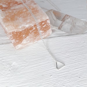 Puede incluir: Un collar de plata con un colgante de tri&aacute;ngulo en una cadena delicada. El collar se muestra sobre una superficie blanca con una roca de sal rosa y un cristal transparente.
