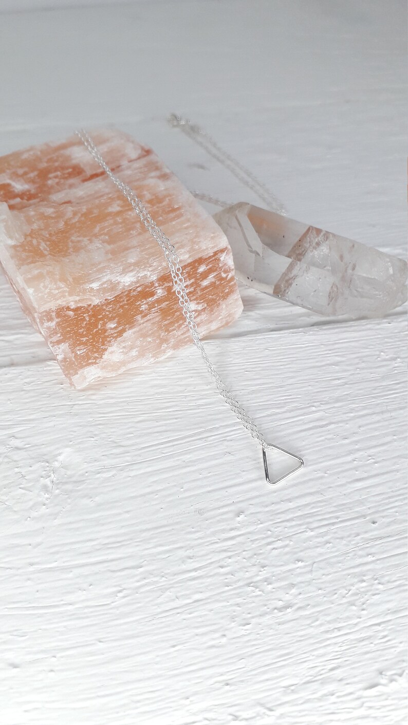 Puede incluir: Un collar de plata con un colgante de tri&aacute;ngulo en una cadena delicada. El collar se muestra sobre una superficie blanca con una roca de sal rosa y un cristal transparente.